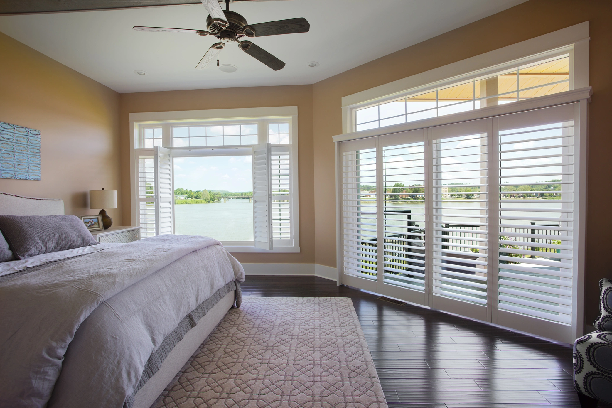 An insulated, airy bedroom in Albuquerque showcases advanced roofing techniques with a large bed, ceiling fan, and windows overlooking a serene lake, all adorned in a cozy beige and brown decor.